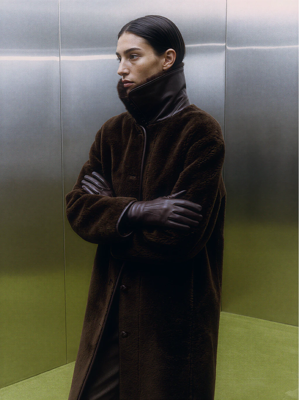 Editorial fashion portrait in a dark coat against brushed metal and green walls.