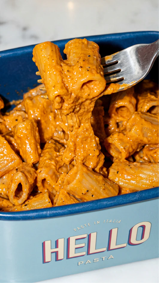 Fork lifting creamy pasta from a blue Hello Pasta takeaway box.