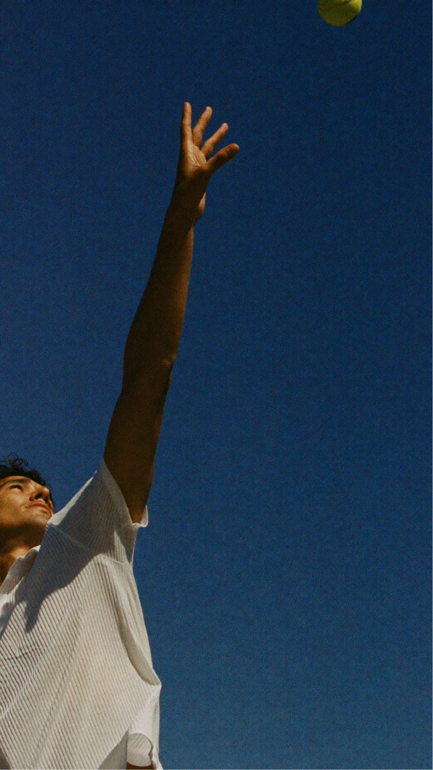 Tennis player reaching upward against a deep blue sky.