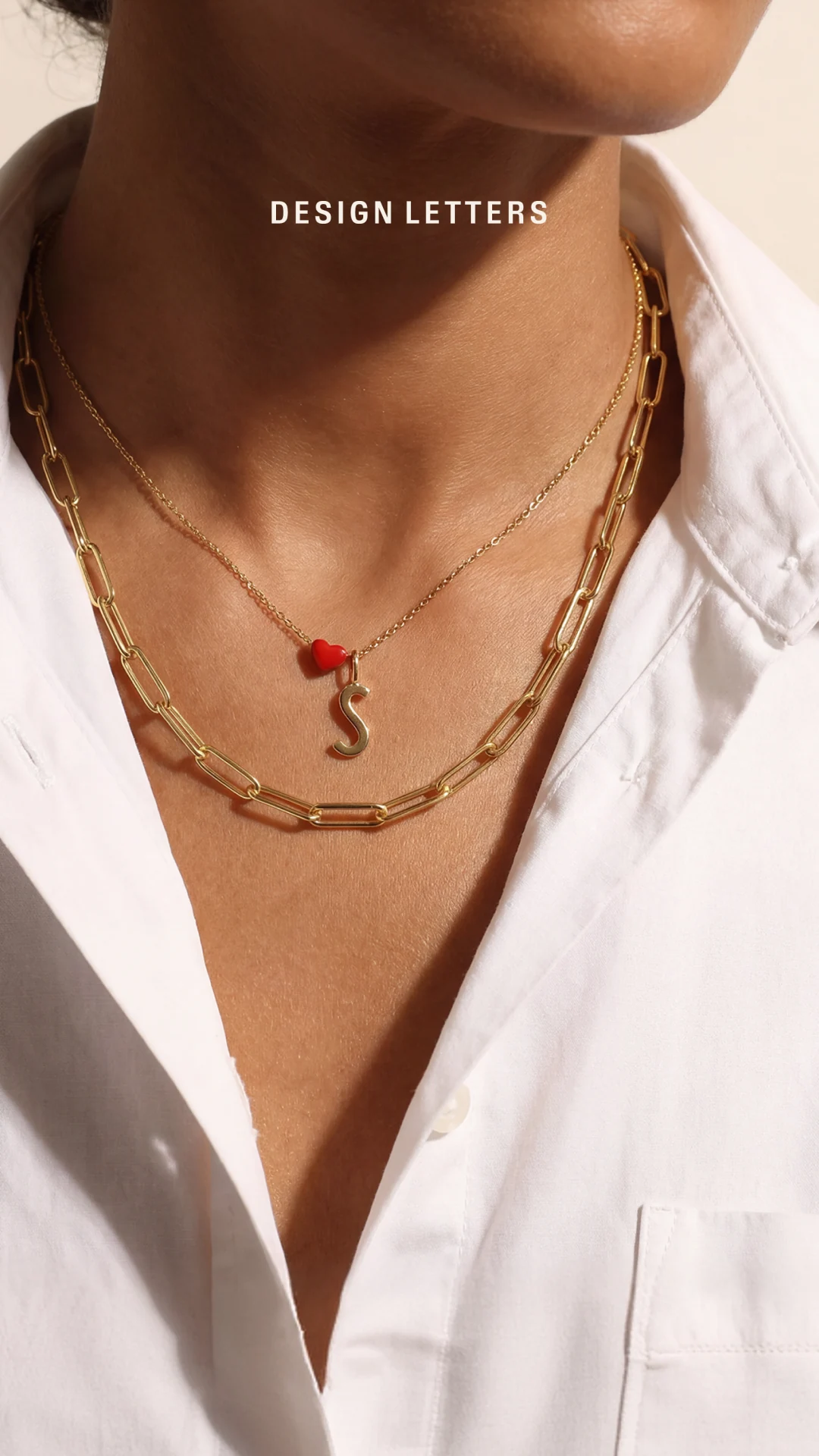 Design Letters gold necklaces layered over a white shirt, with a red heart and letter pendant.